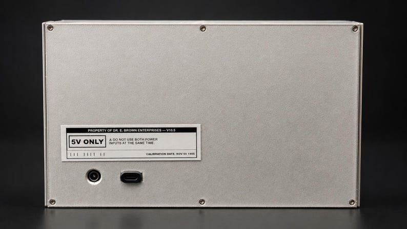May include: A rectangular silver metal electronic device enclosure with four corner screws. A white label reads: PROPERTY OF DR. E. BROWN ENTERPRISES &mdash; V10.5, 5V ONLY, A DO NOT USE BOTH POWER INPUTS AT THE SAME TIME, CALIBRATION DATE: NOV 05 1955. Two power ports are visible.