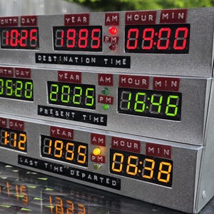 May include: A silver-colored electronic time display unit with three horizontal rows of digital LED screens. The top row displays red text, the middle row green, and the bottom row orange. Labels include DESTINATION TIME, PRESENT TIME, and LAST TIME DEPARTED.