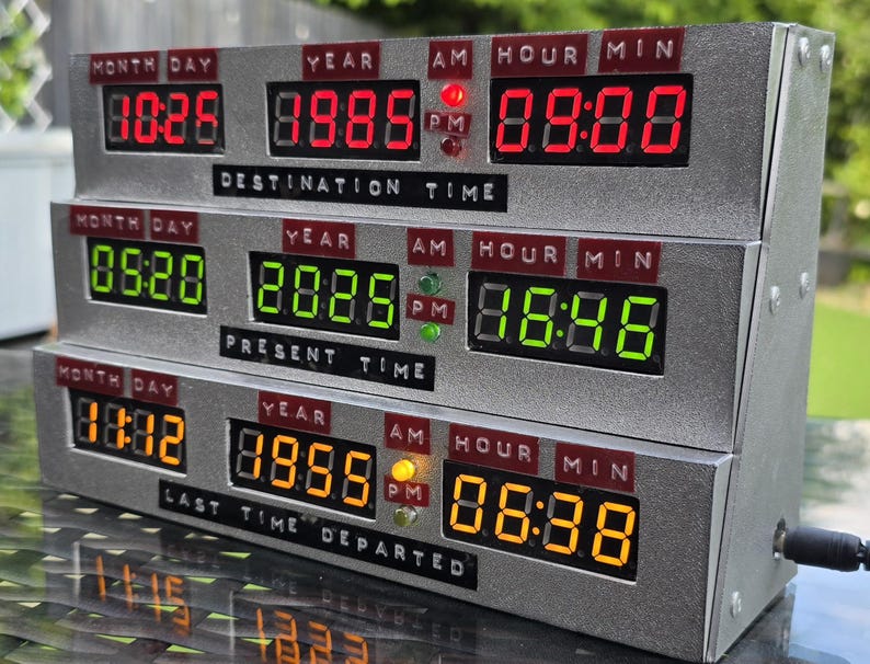 May include: A silver-colored electronic time display unit with three horizontal rows of digital LED screens. The top row displays red text, the middle row green, and the bottom row orange. Labels include DESTINATION TIME, PRESENT TIME, and LAST TIME DEPARTED.