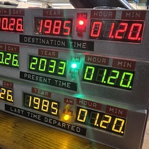 May include: A three-tiered electronic time display device with digital LED readouts. The top row shows red text for Destination Time, the middle row shows green text for Present Time, and the bottom row shows yellow text for Last Time Departed.