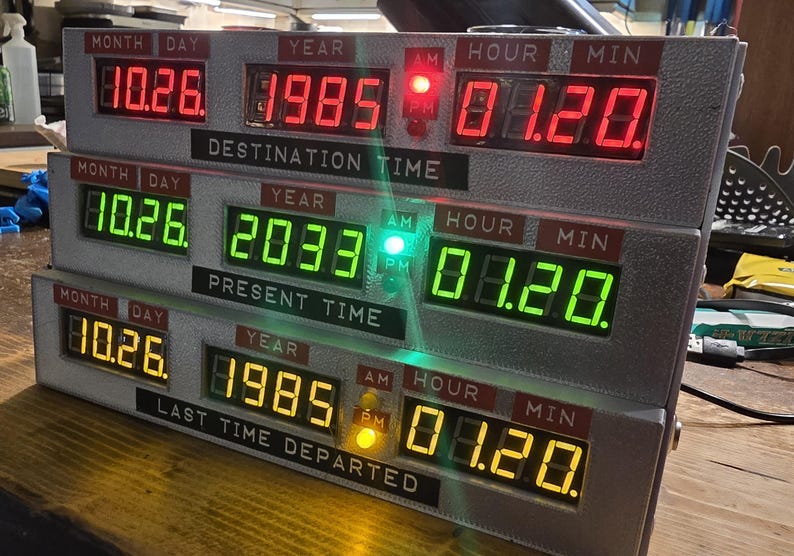 May include: A three-tiered electronic time display device with digital LED readouts. The top row shows red text for Destination Time, the middle row shows green text for Present Time, and the bottom row shows yellow text for Last Time Departed.