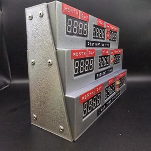 May include: A silver-colored time display unit featuring three stacked digital panels labeled Destination Time, Present Time, and Last Time Departed. Each panel includes red labels for Month, Day, Year, and Time, with black digital display screens.