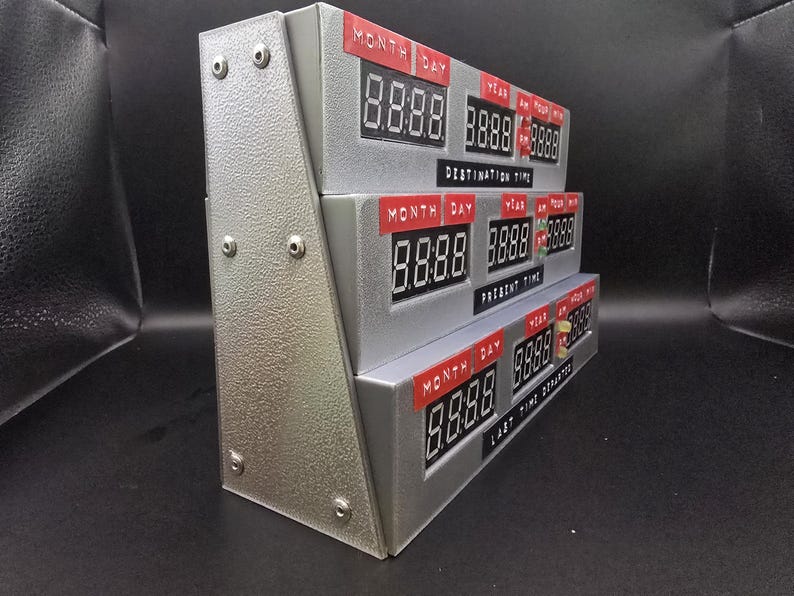 May include: A silver-colored time display unit featuring three stacked digital panels labeled Destination Time, Present Time, and Last Time Departed. Each panel includes red labels for Month, Day, Year, and Time, with black digital display screens.