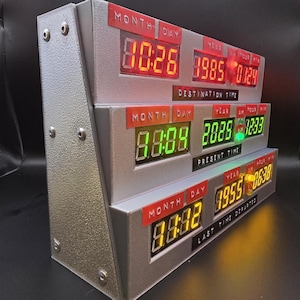 May include: A silver, three-tiered electronic time display unit featuring digital LED readouts in red, green, and yellow. The labels Destination Time, Present Time, and Last Time Departed are printed below each respective row of digital numbers.