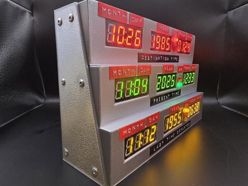 May include: A silver, three-tiered electronic time display unit featuring digital LED readouts in red, green, and yellow. The labels Destination Time, Present Time, and Last Time Departed are printed below each respective row of digital numbers.