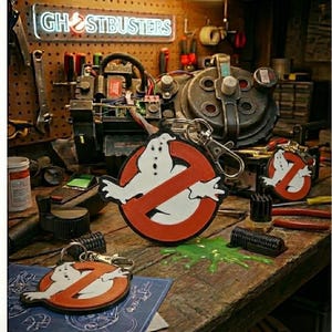 May include: Keyrings featuring the Ghostbusters logo, a white ghost inside a red circle with a slash. The keyrings are on a wooden surface with a green slime-like substance. The background includes a Ghostbusters sign and a proton pack.
