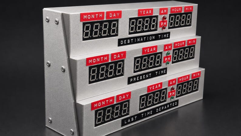 May include: A silver-colored tiered digital clock featuring three separate displays labeled Destination Time, Present Time, and Last Time Departed. Each section includes red labels for Month, Day, Year, AM, PM, Hour, and Min with black digital screens.