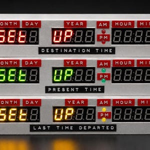 May include: A silver rectangular time display unit with three horizontal rows of digital LED readouts. Each row features red labels for MONTH, DAY, YEAR, AM, PM, HOUR, and MIN. The top row displays red text, the middle green, and the bottom yellow.