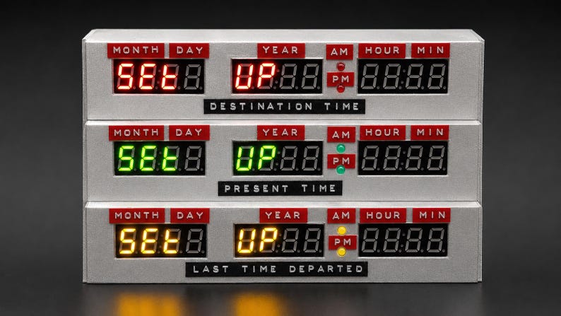 May include: A silver rectangular time display unit with three horizontal rows of digital LED readouts. Each row features red labels for MONTH, DAY, YEAR, AM, PM, HOUR, and MIN. The top row displays red text, the middle green, and the bottom yellow.
