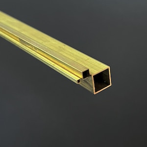 Puede incluir: Primer plano de un tubo cuadrado de latón con un recorte rectangular. El tubo tiene un acabado dorado pulido sobre un fondo gris oscuro. El extremo cuadrado es visible.