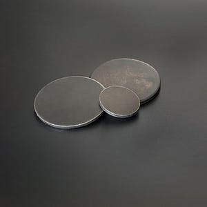 Op de afbeelding: Drie ronde metalen onderzetters in verschillende maten. De onderzetters zijn donkergrijs van kleur met een licht getextureerd oppervlak.