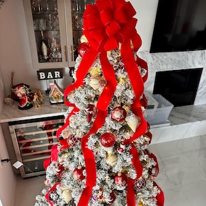 May include: A flocked Christmas tree decorated with red velvet ribbon and ornaments. The tree is topped with a large red velvet bow.