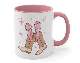 May include: White ceramic mug with a pink rim and handle. The mug features a cartoon illustration of two brown cowboy boots tied together with a pink bow. The background is white with pink stars.