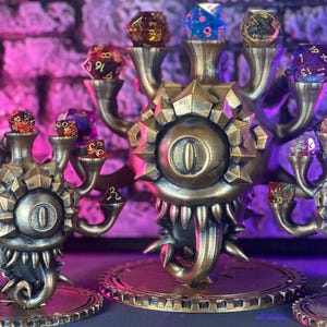 Pode incluir: Suportes de dados com tema de fantasia, na cor bronze, em forma de cabeça de monstro com vários braços, cada um segurando um dado de várias faces. Os dados são de várias cores, incluindo vermelho, azul e transparente. Os suportes assentam sobre uma base circular.