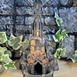 Könnte beinhalten: Ein detailliertes Modell des Dunklen Turms aus Der Herr der Ringe, in dunkelgrau gefertigt mit einem leuchtend orangefarbenen Licht im Inneren. Der Turm ist auf einem felsigen Sockel gebaut und hat ein Tor am Boden.