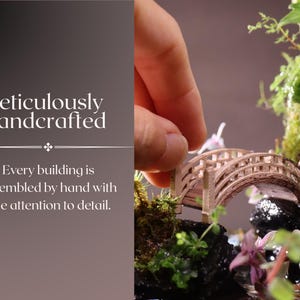 May include: A miniature wooden bridge with intricate details, surrounded by lush greenery and a small waterfall. The bridge is being carefully placed by a hand, highlighting the meticulous craftsmanship of this miniature garden.
