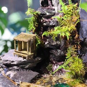 Op de afbeelding: Een miniatuur zen-tuin met een waterval, een klein houten huisje en een houten boot. De tuin is versierd met mos, stenen en kiezelsteentjes.