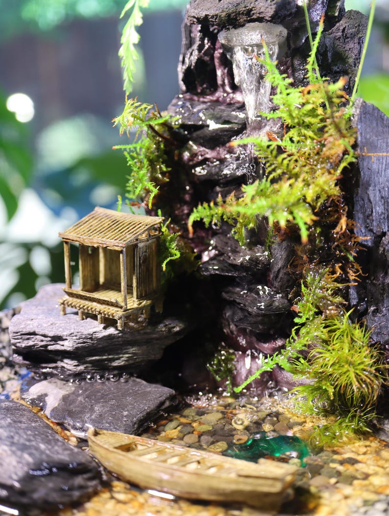Op de afbeelding: Een miniatuur zen-tuin met een waterval, een klein houten huisje en een houten boot. De tuin is versierd met mos, stenen en kiezelsteentjes.