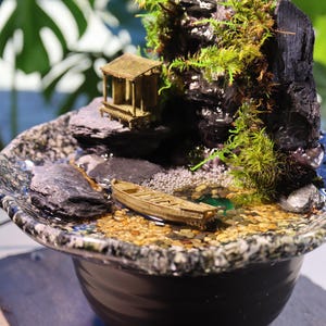 Op de afbeelding: Een miniatuur zen-tuin met een waterval, een klein houten huisje, een boot en groen mos. De tuin is geplaatst in een zwarte keramische pot met een gevlekt oppervlak.
