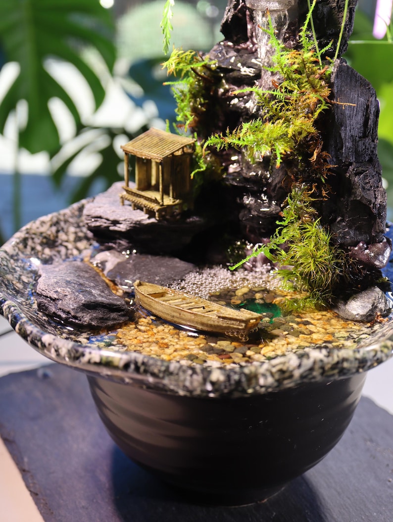 Op de afbeelding: Een miniatuur zen-tuin met een waterval, een klein houten huisje, een boot en groen mos. De tuin is geplaatst in een zwarte keramische pot met een gevlekt oppervlak.