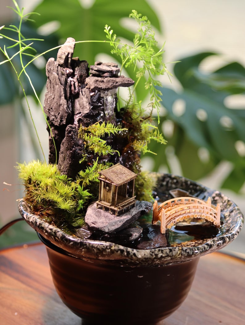 Op de afbeelding: Een miniatuur zen-tuin met een waterval, een klein huis, een brug en mos dat op rotsen groeit in een aardewerken pot.