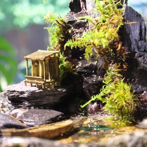 Op de afbeelding: Een miniatuur tafelfontein met een waterval die naar beneden stroomt over een rotsachtige berg, een klein houten huisje en weelderig groen mos. De fontein is geplaatst in een bruine keramische pot.