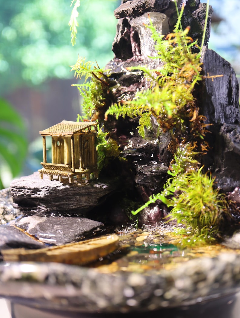 Op de afbeelding: Een miniatuur tafelfontein met een waterval die naar beneden stroomt over een rotsachtige berg, een klein houten huisje en weelderig groen mos. De fontein is geplaatst in een bruine keramische pot.