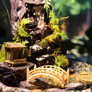 Op de afbeelding: Een miniatuur zen-tuin met een klein houten huisje, een brug en een waterval. De tuin is geplaatst in een bruine keramische schaal met een mosgroene basis.