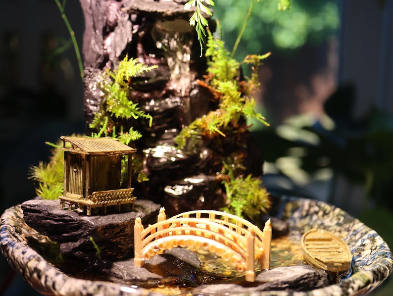 Op de afbeelding: Een miniatuur zen-tuin met een klein houten huisje, een brug en een waterval. De tuin is geplaatst in een bruine keramische schaal met een mosgroene basis.