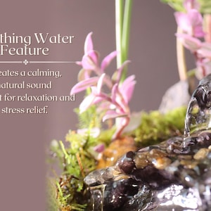 May include: A small tabletop water fountain with a cascading waterfall effect. The fountain is surrounded by green moss and pink flowers. The text "Soothing Water Feature" is displayed above the fountain. The text "Creates a calming, natural sound perfect for relaxation and stress relief." is displayed below the fountain.
