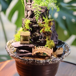 Op de afbeelding: Een miniatuur zen-tuin met een waterval, een brug en een klein huis in een bruine keramische pot. De tuin is versierd met mos, stenen en een klein bootje.