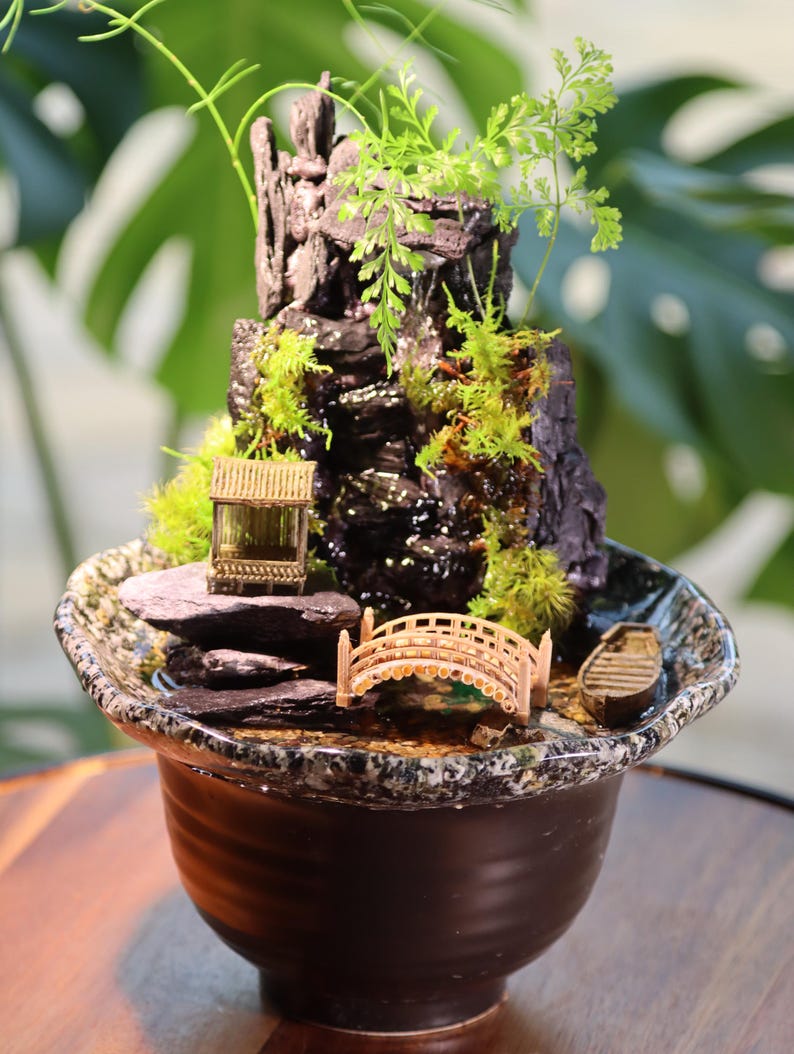 Op de afbeelding: Een miniatuur zen-tuin met een waterval, een brug en een klein huis in een bruine keramische pot. De tuin is versierd met mos, stenen en een klein bootje.