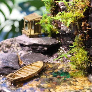 Op de afbeelding: Een miniatuur houten bootje drijft in een klein vijvertje omringd door stenen en mos. De vijver is onderdeel van een zen-tuin op tafel met een miniatuur houten huisje en een waterval.