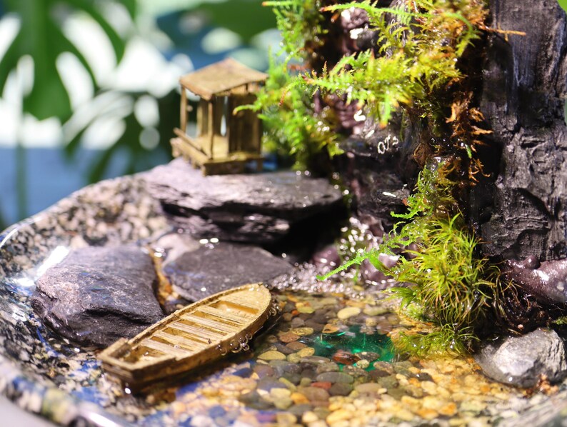 Op de afbeelding: Een miniatuur houten bootje drijft in een klein vijvertje omringd door stenen en mos. De vijver is onderdeel van een zen-tuin op tafel met een miniatuur houten huisje en een waterval.
