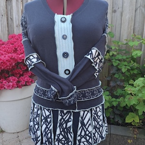 Op de afbeelding: Marineblau vestjurk met V-hals en knoopsluiting. Het heeft een lichtblauw middenpaneel en decoratieve mouwen en rok met patroon. De rok is geplooid met een wit en marineblauw geometrisch patroon. Getoond op een mannequin.