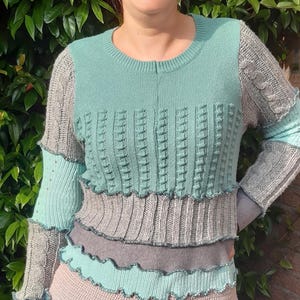 Könnte beinhalten: Ein tealblauer, grauer und rosafarbener Strickpullover mit einem Rüschen-Saum. Der Pullover besteht aus verschiedenen Strickstoffen und hat einen geschichteten Look.