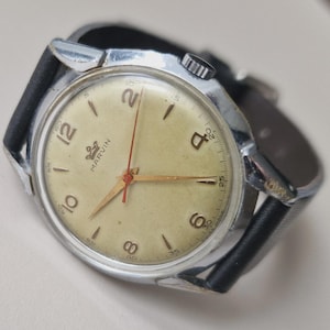 Può includere: Orologio da polso Marvin vintage con cassa color argento e quadrante color crema. L'orologio presenta numeri neri, lancette dorate e lancetta dei secondi rossa. Il cinturino in pelle nera completa il design classico. Il quadrante mostra il logo Marvin.