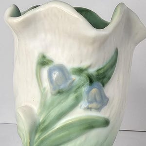 May include: A white ceramic vase with a ruffled rim and a floral design. The vase features blue bell-shaped flowers and green leaves. The base of the vase is also green.