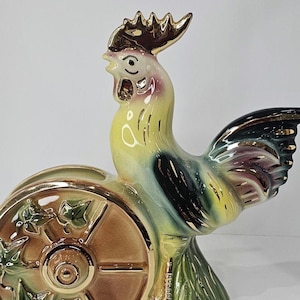 May include: A ceramic rooster figurine with a decorative wheel. The rooster is yellow, green, and purple with gold accents. The wheel is brown with green ivy detailing and gold trim. The figurine is on a green base.
