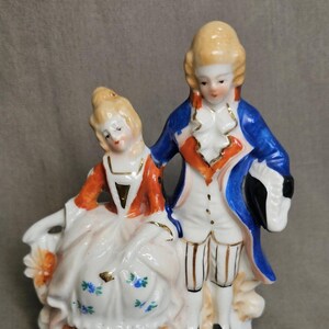 May include: A porcelain figurine depicting a couple in 18th-century attire. The man wears a blue coat and white breeches, while the woman is in a white dress with orange accents. The base is white with a decorative border.