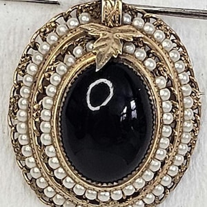 May include: Oval gold-tone brooch with a large black center stone. The brooch is surrounded by small, white faux pearls and a decorative gold filigree border. A gold leaf accent is at the top.