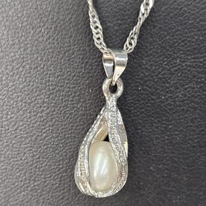 Peut inclure: Un collier en cha&icirc;ne argent avec un pendentif en forme de cage argent&eacute;e contenant une perle blanche.