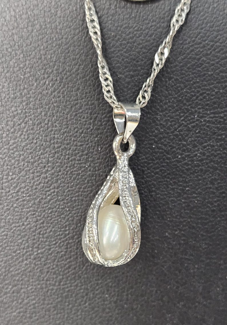 Peut inclure: Un collier en cha&icirc;ne argent avec un pendentif en forme de cage argent&eacute;e contenant une perle blanche.