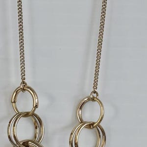 May include: Gold-toned necklace featuring a chain and multiple linked circles. The necklace has a delicate chain and a series of interconnected, large circular links. The jewelry is displayed against a white background.
