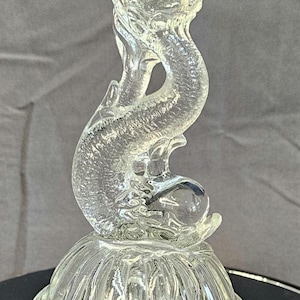 May include: Clear glass candlestick holder with a dolphin design. The candlestick features a base with a scalloped edge, a dolphin-shaped stem, and a flared top. The dolphin's body curves gracefully, holding a glass sphere.