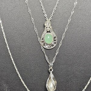 Peut inclure: Un collier argent&eacute; &agrave; trois rangs avec trois pendentifs. Le pendentif du haut pr&eacute;sente une pierre pr&eacute;cieuse verte entour&eacute;e de petites pierres claires. Le pendentif du milieu est en forme de larme avec une perle &agrave; l'int&eacute;rieur. Le pendentif du bas est une simple forme de larme.