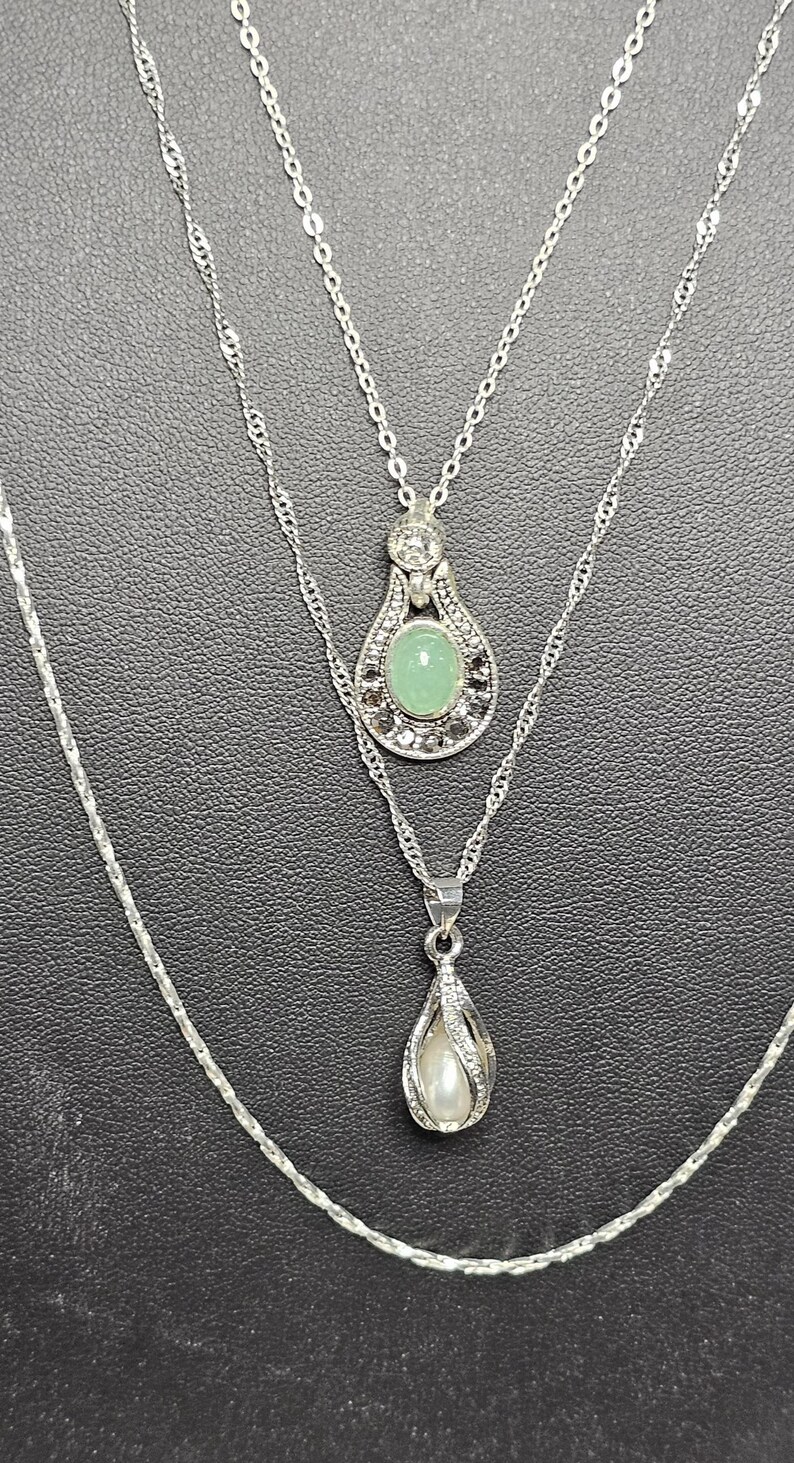 Peut inclure: Un collier argent&eacute; &agrave; trois rangs avec trois pendentifs. Le pendentif du haut pr&eacute;sente une pierre pr&eacute;cieuse verte entour&eacute;e de petites pierres claires. Le pendentif du milieu est en forme de larme avec une perle &agrave; l'int&eacute;rieur. Le pendentif du bas est une simple forme de larme.