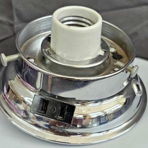 May include: A chrome-plated light fixture base with a white ceramic socket. The base has a circular design with a black outlet and two screws for mounting. The fixture is designed for electrical use.