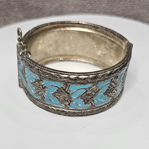 May include: A silver bangle bracelet with a blue enamel design. The bracelet has a floral pattern and is hinged.