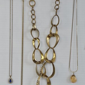 May include: A collection of gold-toned necklaces, each with unique designs. One features a large, linked chain with a crystal-studded pendant. Others include delicate chains with teardrop and oval pendants. The necklaces are displayed against a white background.